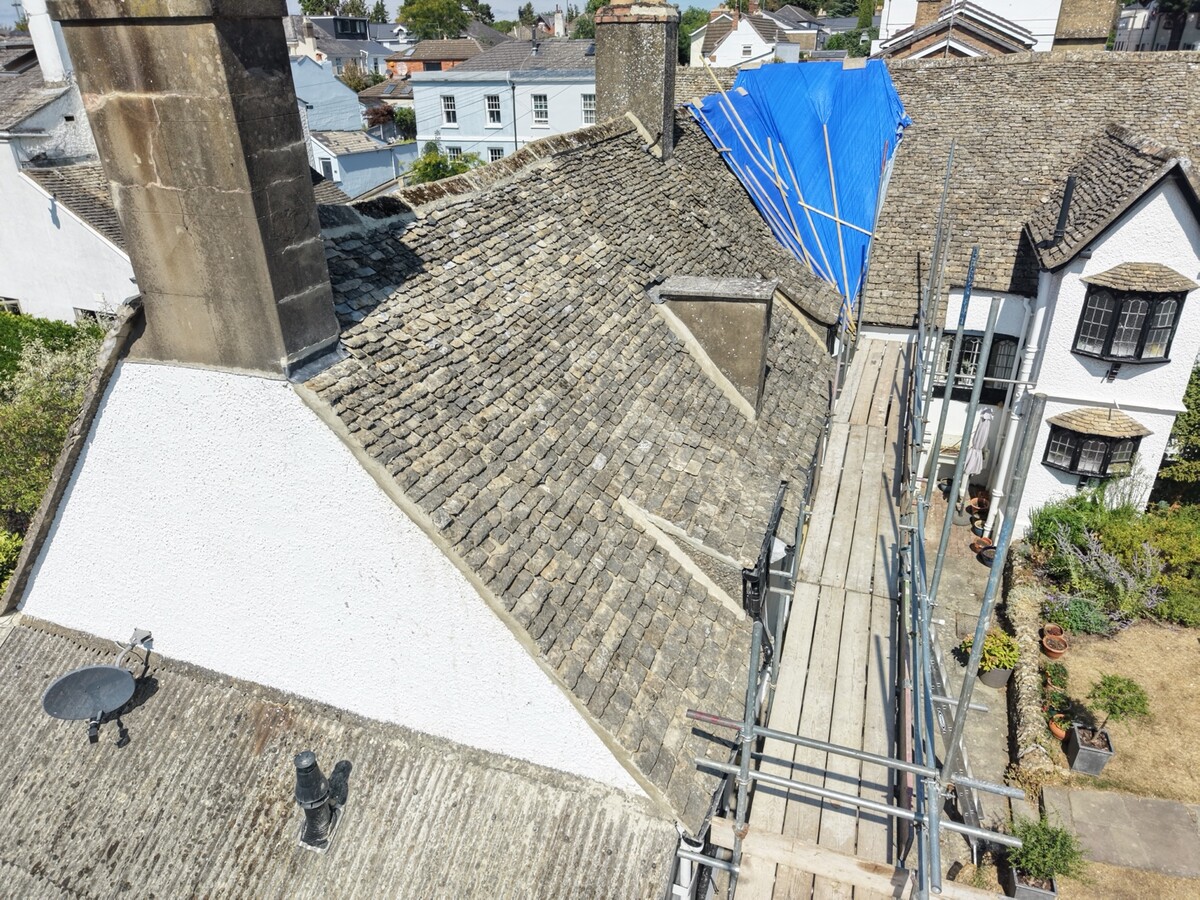 Stone roof and scaffolding with a blue temporary covering on an adjoining roof section at Cudnall Street, Charlton Kings